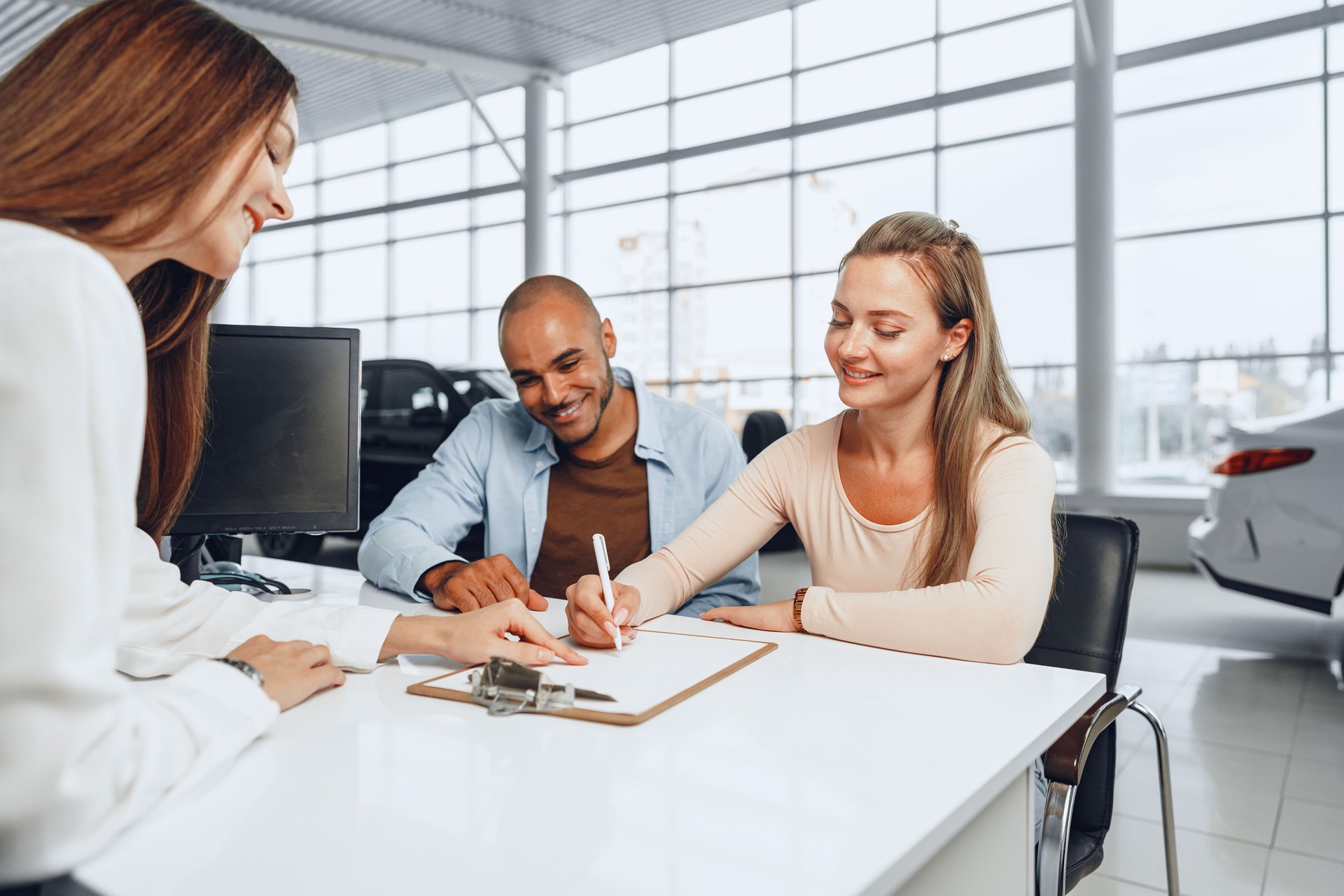 Beautiful young couple signs documents at car dealership showroom